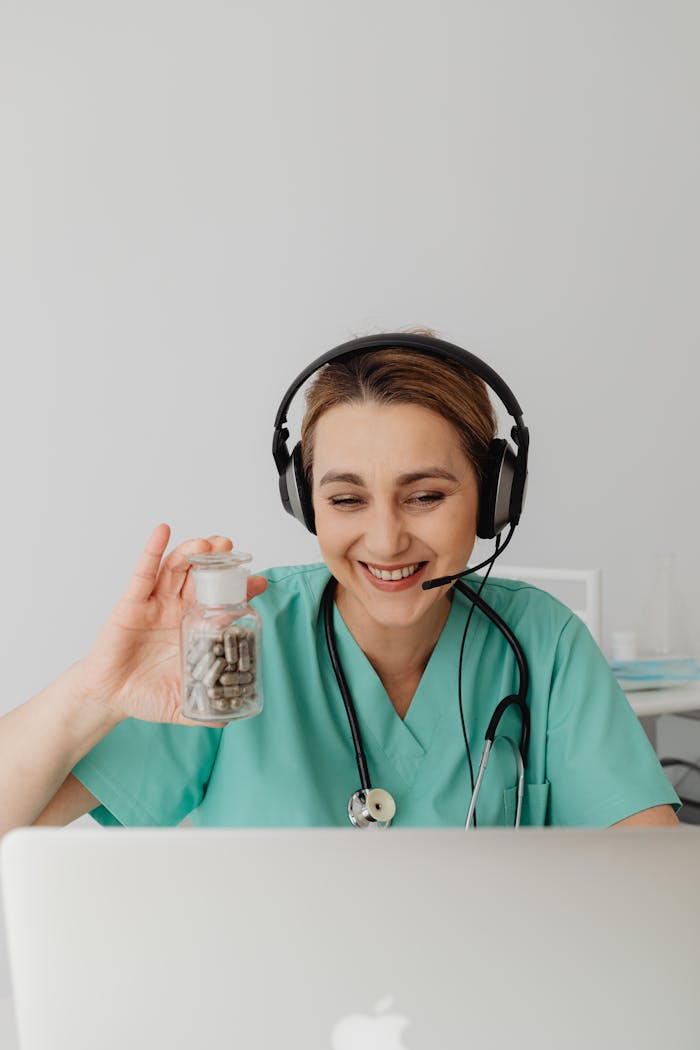 who-we-are Nurse in green scrubs wearing headphones smiles while holding a pill bottle during a video call.